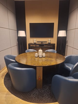 Modern home theater room with round wooden table, blue leather chairs, large TV mounted on wall, tall speakers, and warm ambient lighting